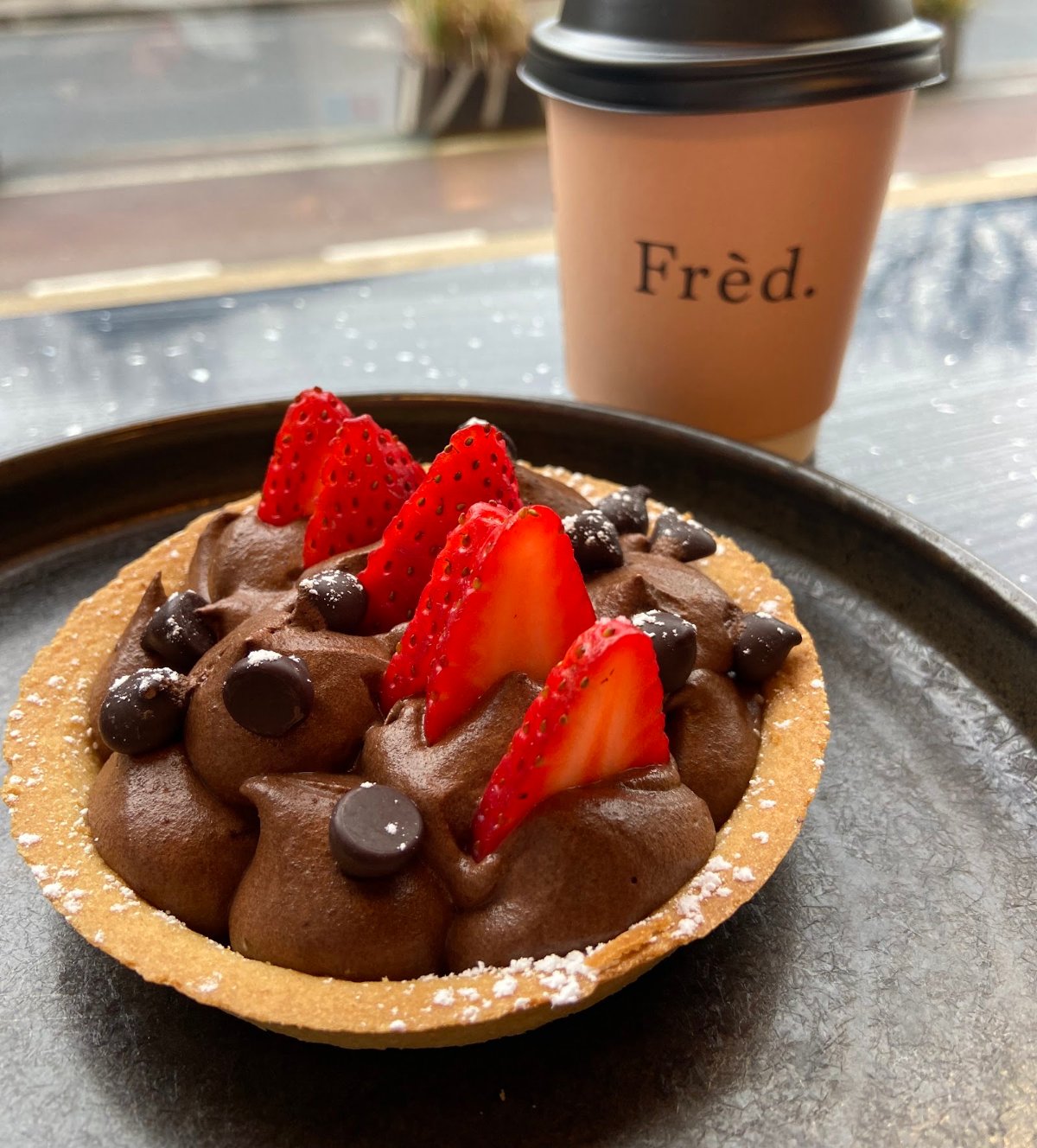 A chocolate tart with strawberries and a Frèd. takeaway cup on a windowsill table, Nassau Street behind.